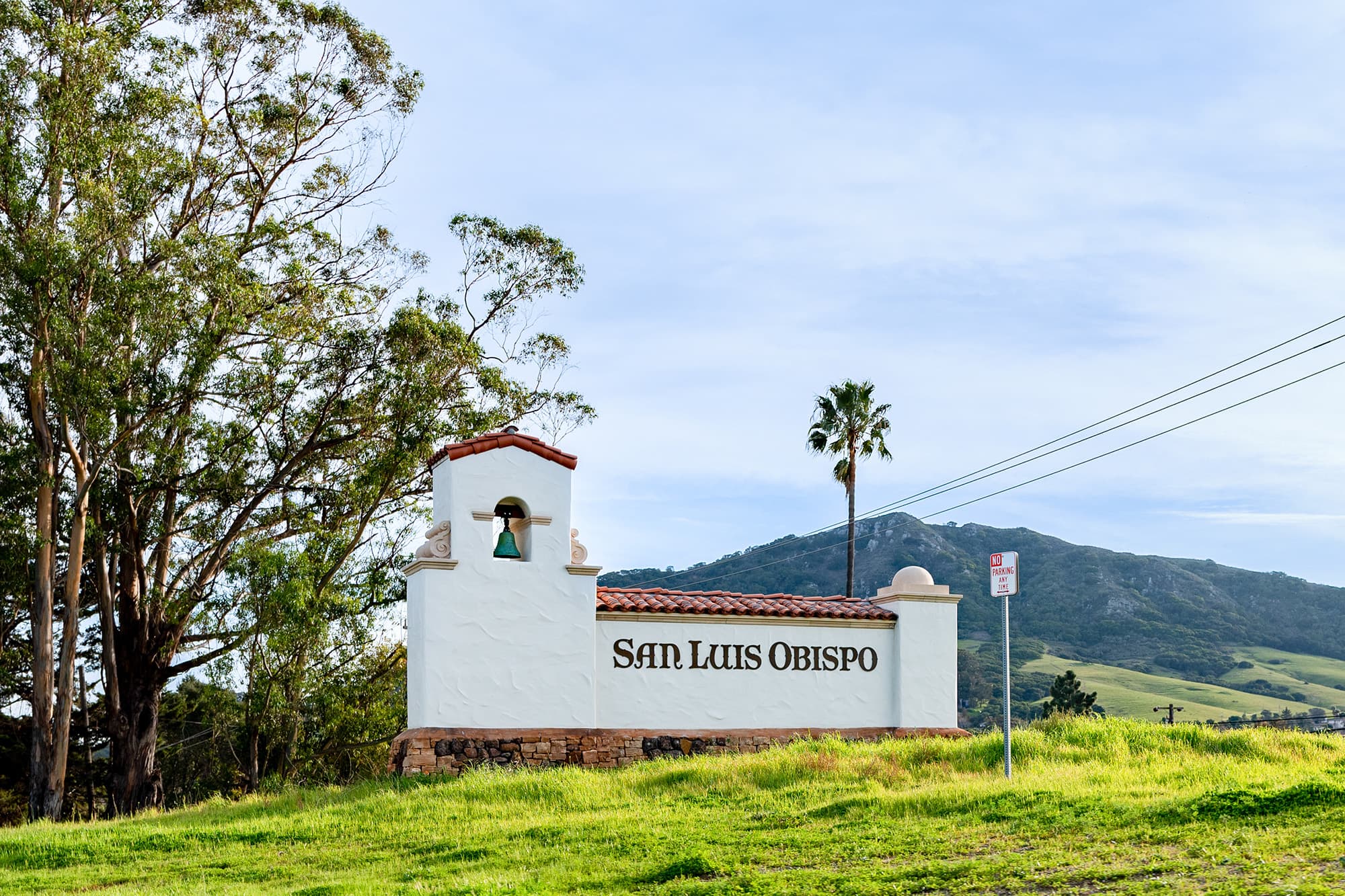 San Luis Obispo sign with a bell tower, green hills, and trees.
