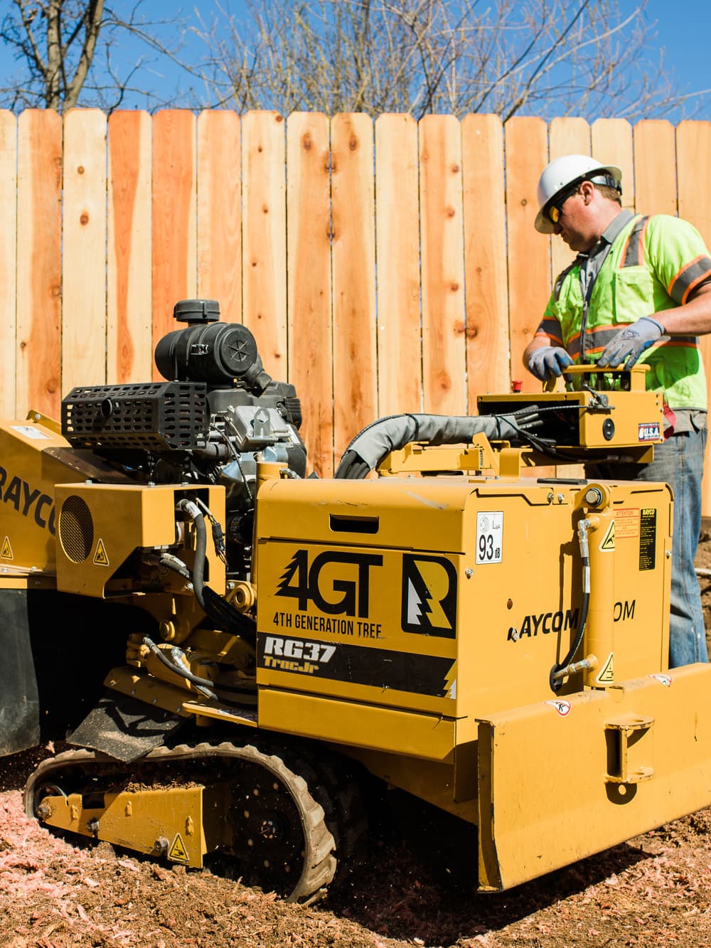 Worker operating a yellow tracked stump grinder beside a wooden fence.