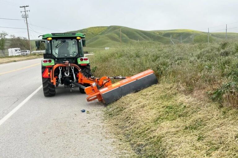 Tractor with a side-mounted mower cutting grass along a roadside.