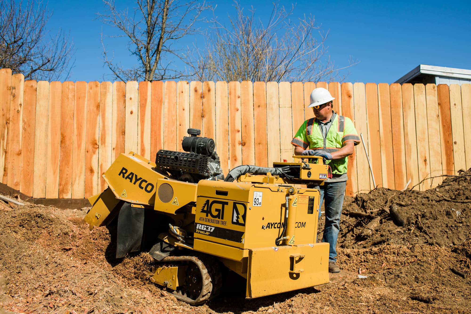 Worker operating a yellow tracked stump grinder beside a wooden fence.