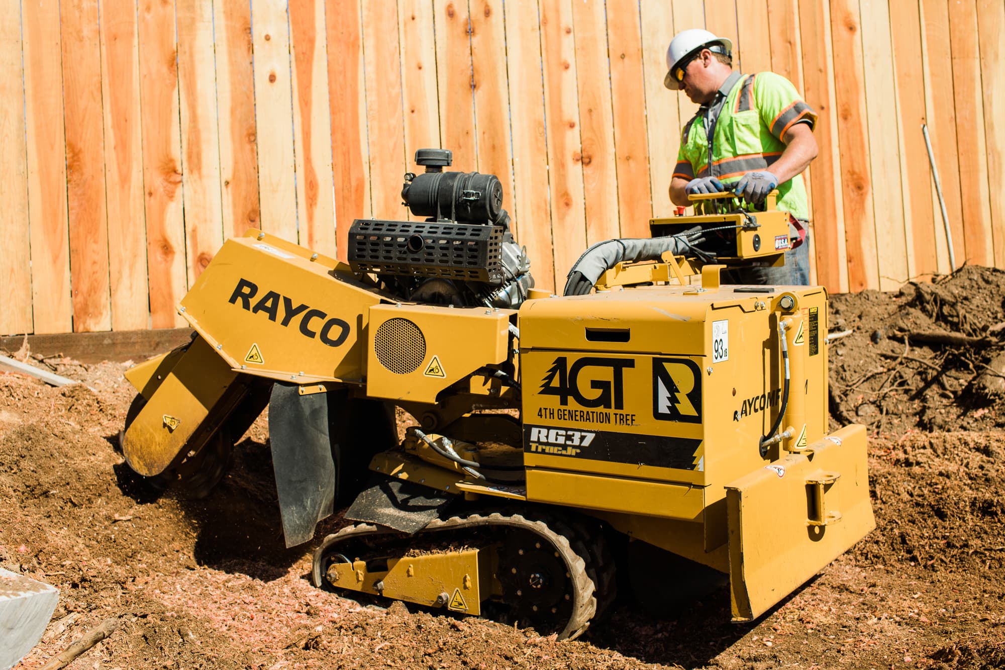 Worker operating a yellow tracked stump grinder beside a wooden fence.